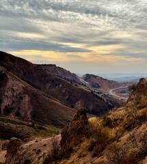 Scenic view of a winding road through arid mountains. Rugged hilly landscape under a dramatic cloudy sky at sunset.
