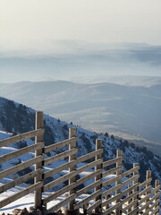 A wooden fence on a snow-covered mountainside overlooking a vast, hazy mountain range.