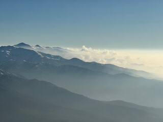 Hazy mountain range with snow-capped peaks. Scenic aerial view of layered mountains and clouds under a clear blue sky.