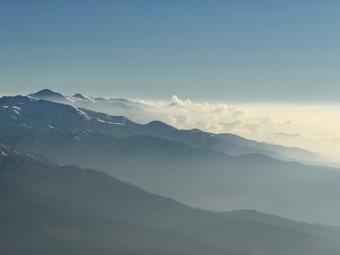 Hazy mountain range with snow-capped peaks. Scenic aerial view of layered mountains and clouds under a clear blue sky.