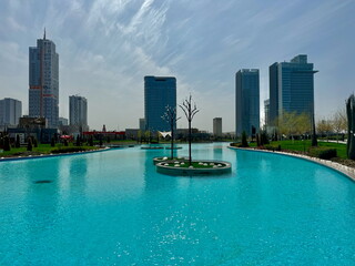 Naklejka premium A modern city park with a bright turquoise river and a skyline of skyscrapers. Urban oasis and public green space.