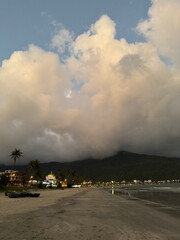 Dramatic clouds loom over a tropical beach at dusk. A coastal town with glowing lights sits at the...