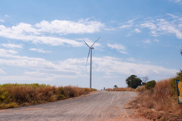 A single wind turbine stands tall against a bright blue sky, surrounded by a rural dirt road and...