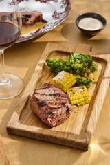 Filet mignon steak on wooden board, bright daylight, overhead composition with grilled corn and...