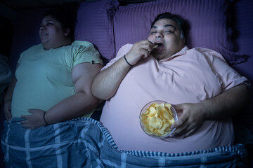 Overweight fat indian husband eating unhealthy fried junk food while lying in bed with her wife. Plus size couple resting together in bedroom. Obesity.