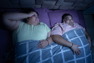 Overweight indian woman unable to sleep as her husband snoring in bed. Plus size couple resting together. Fat woman annoyed with man noisy sound at night while bedtime.