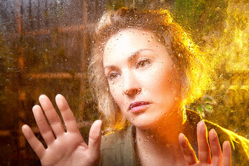 Woman gazes thoughtfully through a rain-soaked window in a moment of reflection during the early...
