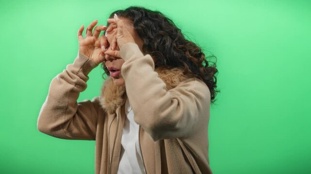 Woman with curly hair makes binoculars gesture against a green background, wearing a beige jacket, suggesting a playful and imaginative setting.