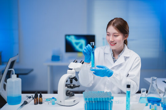 Asian female scientist wearing gloves and lab coat pouring blue liquid substance into test tube while smiling in modern laboratory with microscope and dna structure on computer screen
