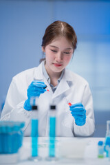 Asian female scientist wearing gloves and lab coat dropping liquid with pipette into test tube...