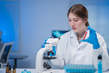 Young scientist wearing lab coat and gloves adjusting microscope and holding blood sample tube in...