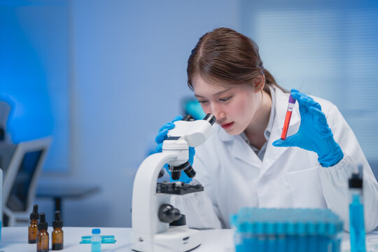 Scientist in a lab coat examines a sample under a microscope, holding a test tube. The laboratory setting is modern, with scientific equipment visible in the background