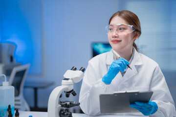 Asian female scientist wearing lab coat and gloves taking notes on clipboard while thinking about...