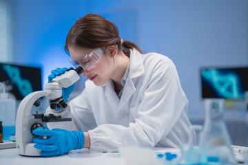 Young female scientist wearing lab coat and gloves using microscope in modern laboratory with dna...