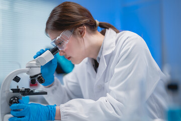 Female scientist in white coat using microscope analyzing sample in laboratory wearing protective...