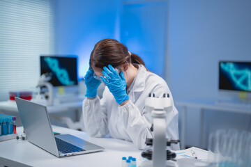 Young scientist in lab coat and gloves sitting at desk, holding her head in hands, expressing...