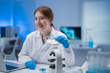 Asian female scientist wearing a lab coat and gloves is smiling while using a microscope in a modern laboratory, with a laptop and test tubes nearby, suggesting advanced scientific research