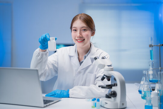Smiling scientist wearing lab coat and gloves showing medicine bottle while sitting at desk with microscope and laptop in laboratory with blue light