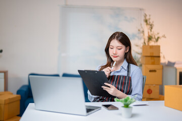 Young Asian SME business woman wearing apron working at home office using laptop checking customer order preparing packing list together with product box parcel