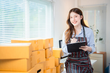 Young asian woman entrepreneur taking notes on clipboard while managing online orders and preparing...