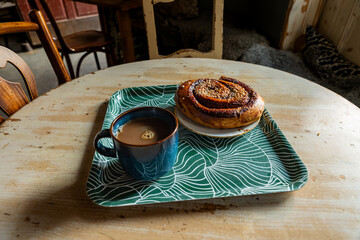 Delicious, sweet, glazed, large cinnamon and cardamom spiced spiral pastry bun, Kanelbulle, topped with syrup and white pearl-sugar sprinkles, with hot cup of coffee in a rustic scene