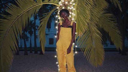 Fashion model wearing yellow jumpsuit standing under palm tree decorated with string lights on tropical beach at night, creating a magical and stylish atmosphere - Powered by Adobe