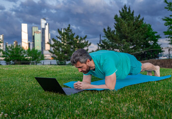 Mature man watching online fitness lesson on laptop. Virtual coach. Plank exercise on mat in the...
