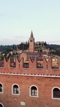 Vertical drone shot frames Verona&rsquo;s Palazzo del Podest&agrave; battlements and the Basilica di San Zeno Maggiore with its bell tower rising on the hill, real time, warm evening light over historic rooftops