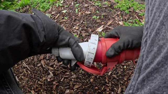 Gloved hands connect heavy-duty industrial power plug into its socket on mulch-covered soil, part of outdoor event or construction setup, red locking arm engaged, real time, close-up, first-person POV