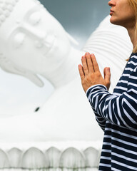 
A young girl of European appearance folds her hands in namaste and meditates in front of a huge white statue of a reclining Buddha.