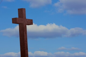 Tall dark wooden cross is standing against blue sky with white clouds in flat design