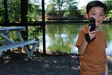 Young Asian boy holds an action camera while standing in a park with picnic tables and trees behind him.