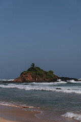 Ocean waves breaking on the shore of the Parrot Rock, a small island with vegetation in Mirissa, Sri Lanka, under a clear sky