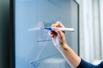 A person writing on a whiteboard with a blue marker in a bright office space