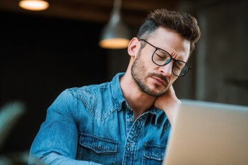 Obraz premium Young male designer wearing glasses and a shirt, sitting at a desk with neck pain, using a computer for work in an office interior.