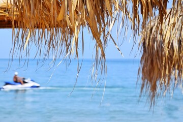 A jet ski is visible through the thatched roof of a beach umbrella. The concept of relaxation and...