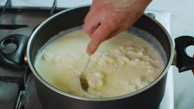 Using a spoon to stir curd in a pot, separating it from whey during the cheese-making process. Close-up shot of traditional homemade dairy preparation