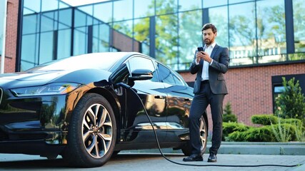Stylish businessman confidently charges modern electric car at urban charging station. Investor attentively uses smartphone waiting. Scene represents sustainable transportation business connectivity. - Powered by Adobe