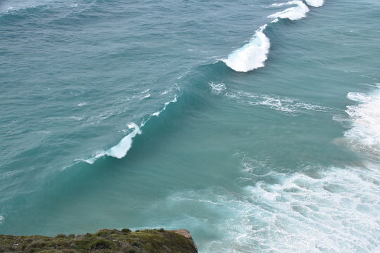 A powerful turquoise wave crashes onto the shore, captured in stunning detail from above. The seafoam swirls in vibrant white against the crystal-clear blue water