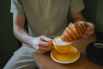 Close-up of a man sitting in a cafe dipping a croissant in a cup of coffee.