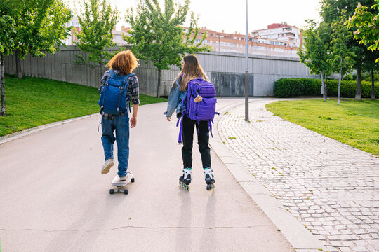 Two friends enjoying a sunny day skating and longboarding in a city park, wearing backpacks and casual clothes
