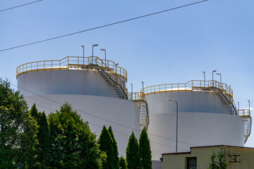 Two large fuel storage tanks with railings and stairs, surrounded by greenery and industrial...