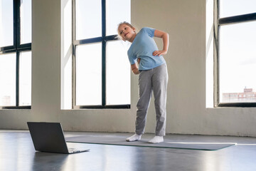 Girl practicing yoga indoors while looking at laptop in a sunlit space.