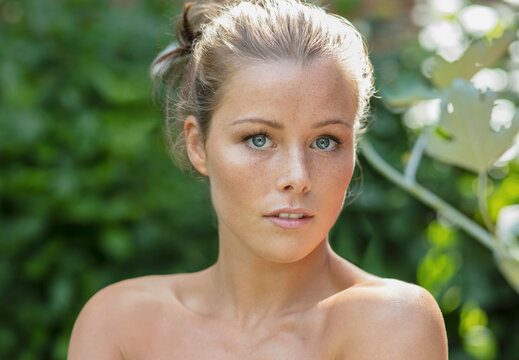 Portrait of beautiful, wide-eyed young woman with bare shoulders and freckles in summer garden
