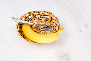 Ripe banana and fork in a small wicker basket on a white wooden table.