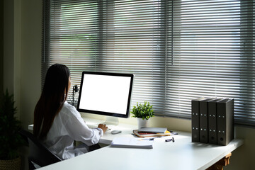 Young woman in a white shirt working on a blank screen desktop in front of window blinds