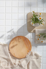 Bathroom flatlay with beige fabric, round wood trays, and flower decor on square white tiles. A...