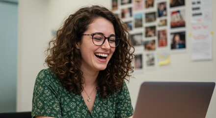 Young woman with curly hair and glasses laughing joyfully while looking at her laptop screen in an office setting