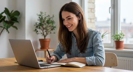 Young woman smiling while working on a laptop and writing notes in a notebook at a wooden desk