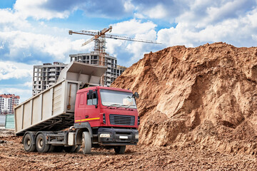 Dump truck unloading soil at a construction site under a cloudy sky in an urban area